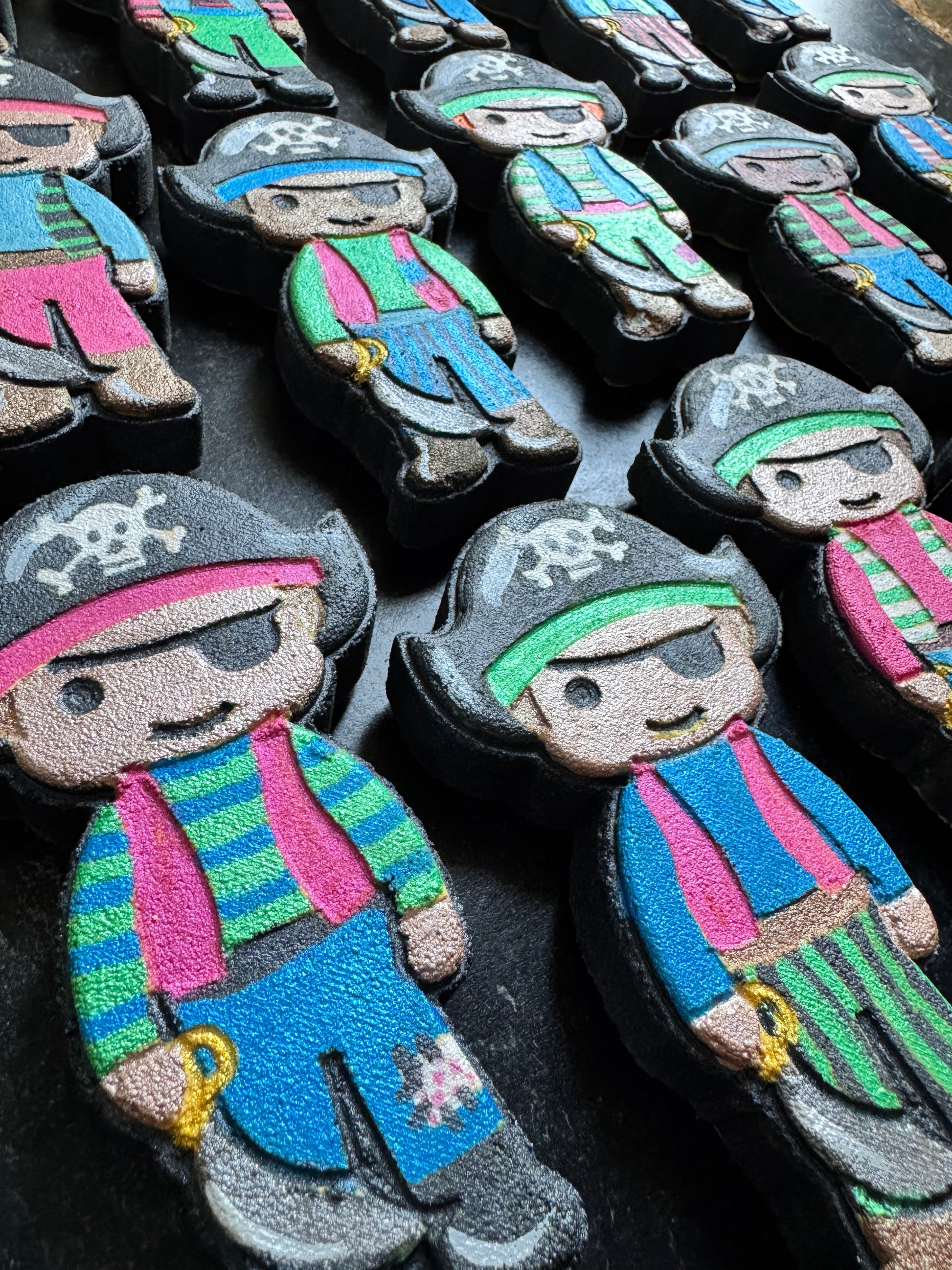 A close-up of multiple pirate-shaped items arranged on a dark surface. Each item is decorated with colorful pirate clothing and accessories, including pink and green striped shirts, blue pants, black hats, and small yellow accents.
