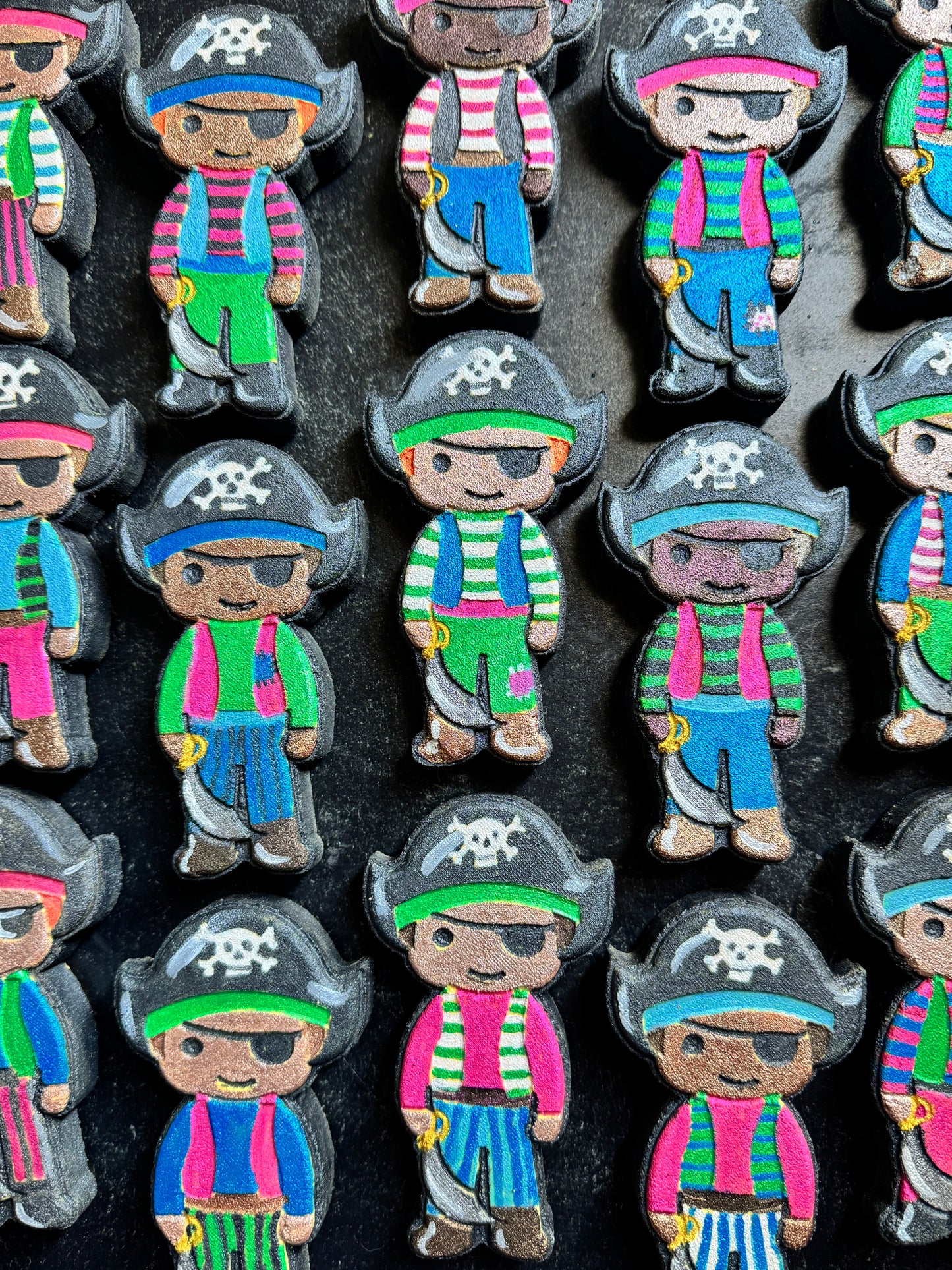 A close-up of multiple hand-painted pirate-shaped cookies arranged in rows on a dark background. Each cookie features a cartoon pirate with a black hat, eyepatch, colorful striped shirt, blue pants, and a small yellow treasure chest.
