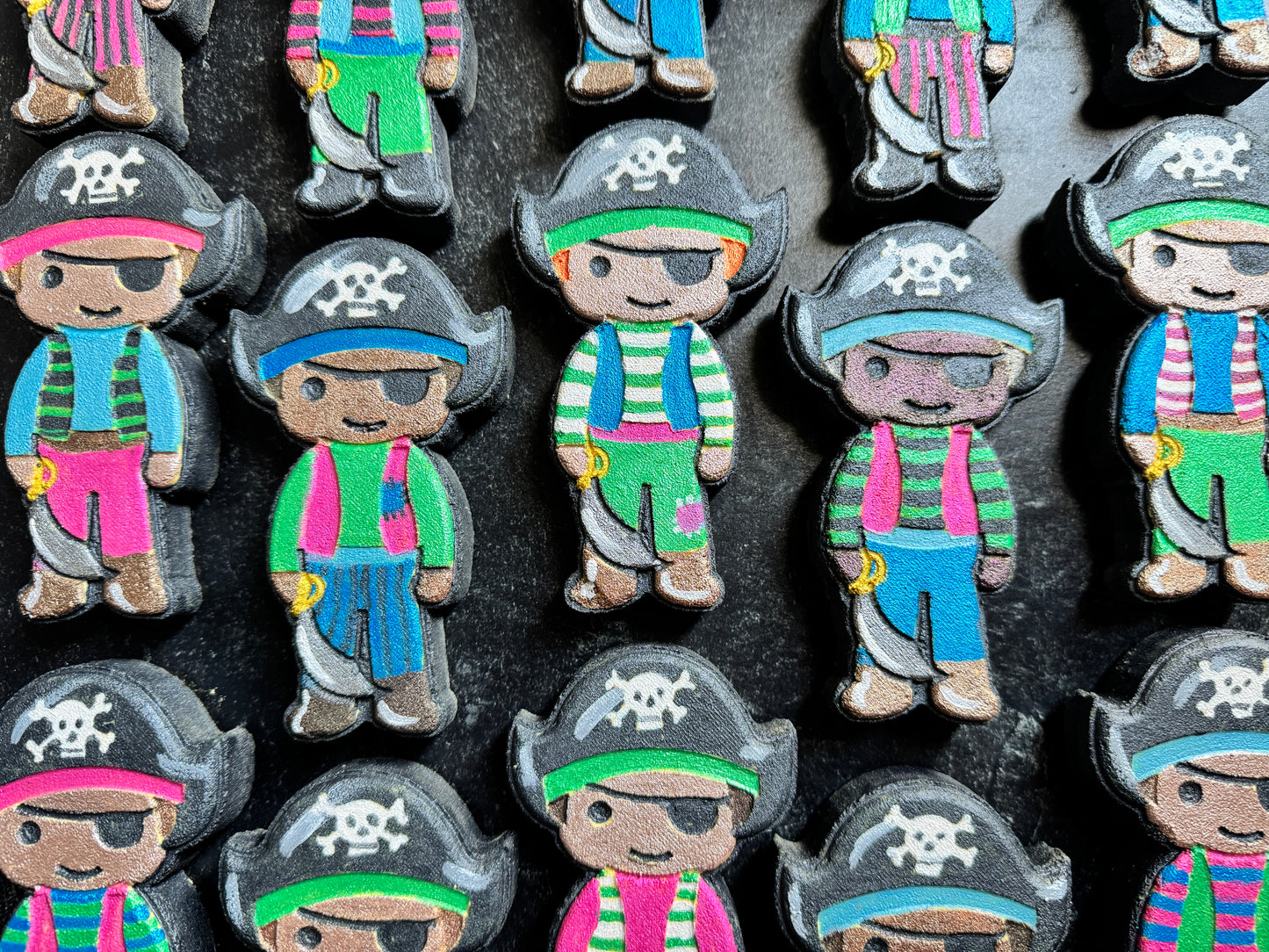 A close-up of multiple pirate-shaped craft charms or stickers arranged on a dark surface. Each character wears a black pirate hat with a white skull-and-crossbones emblem, an eyepatch, and colorful striped clothing in pink, green, blue, and red, with some holding a gold sword or pistol.